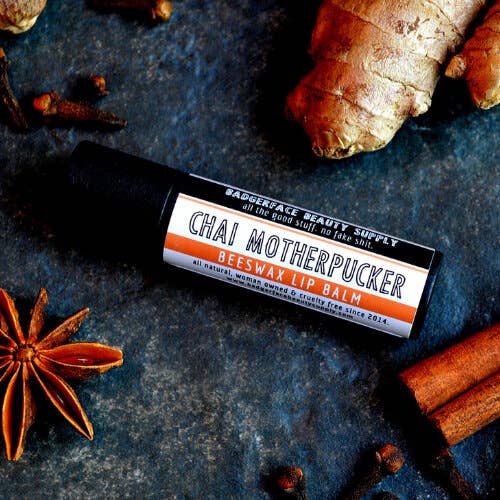 An earthy photo showing ginger and cinnamon sticks with a tube of Chai Motherpucker lip balm. The label text shows, "Pucker up, bitches!"