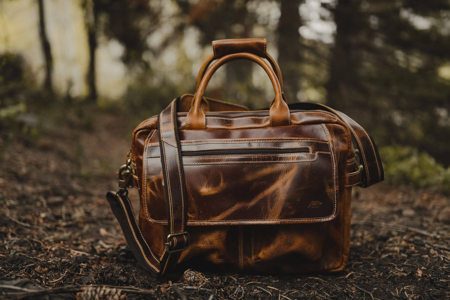 Leather pilot bag sitting on the ground outside in a wooded environment showing the handles and shoulder strap.