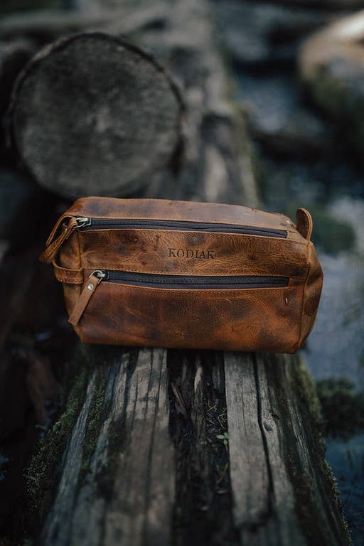 Brown leather dopp kit on a wooden log with a blurred natural background